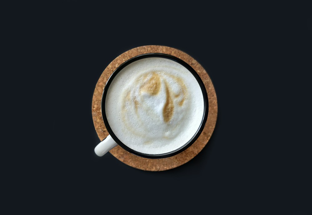 Overhead photograph of a foamy drink in a white cup on a cork coaster against a dark background