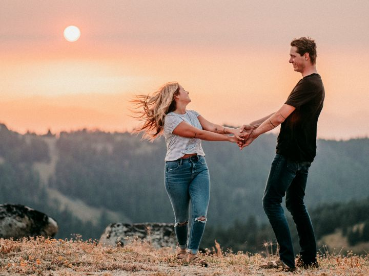 A couple hold both hands in an open landscape with a low sun, distant trees and warm sky.