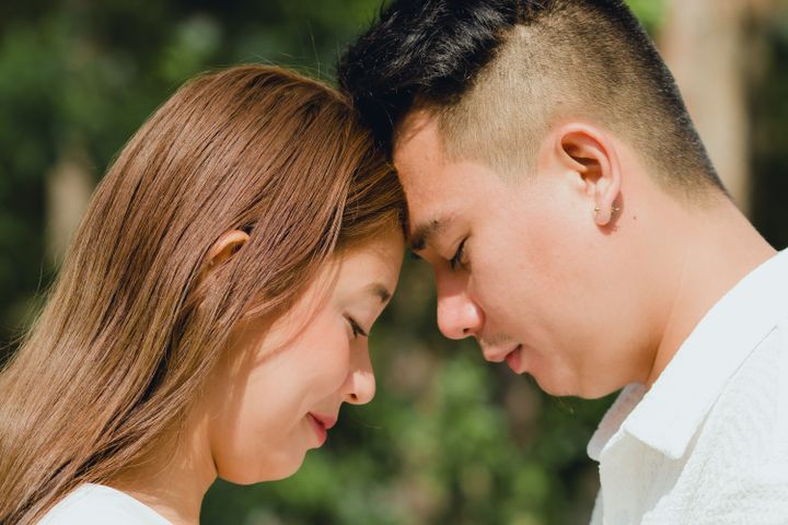 A man and woman touch foreheads with their eyes closed against softly blurred green foliage.
