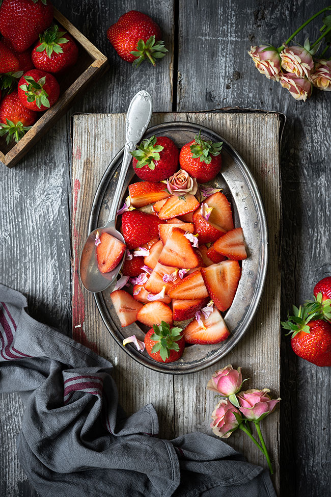 Overhead strawberries on an oval platter with a spoon, roses and gray cloth