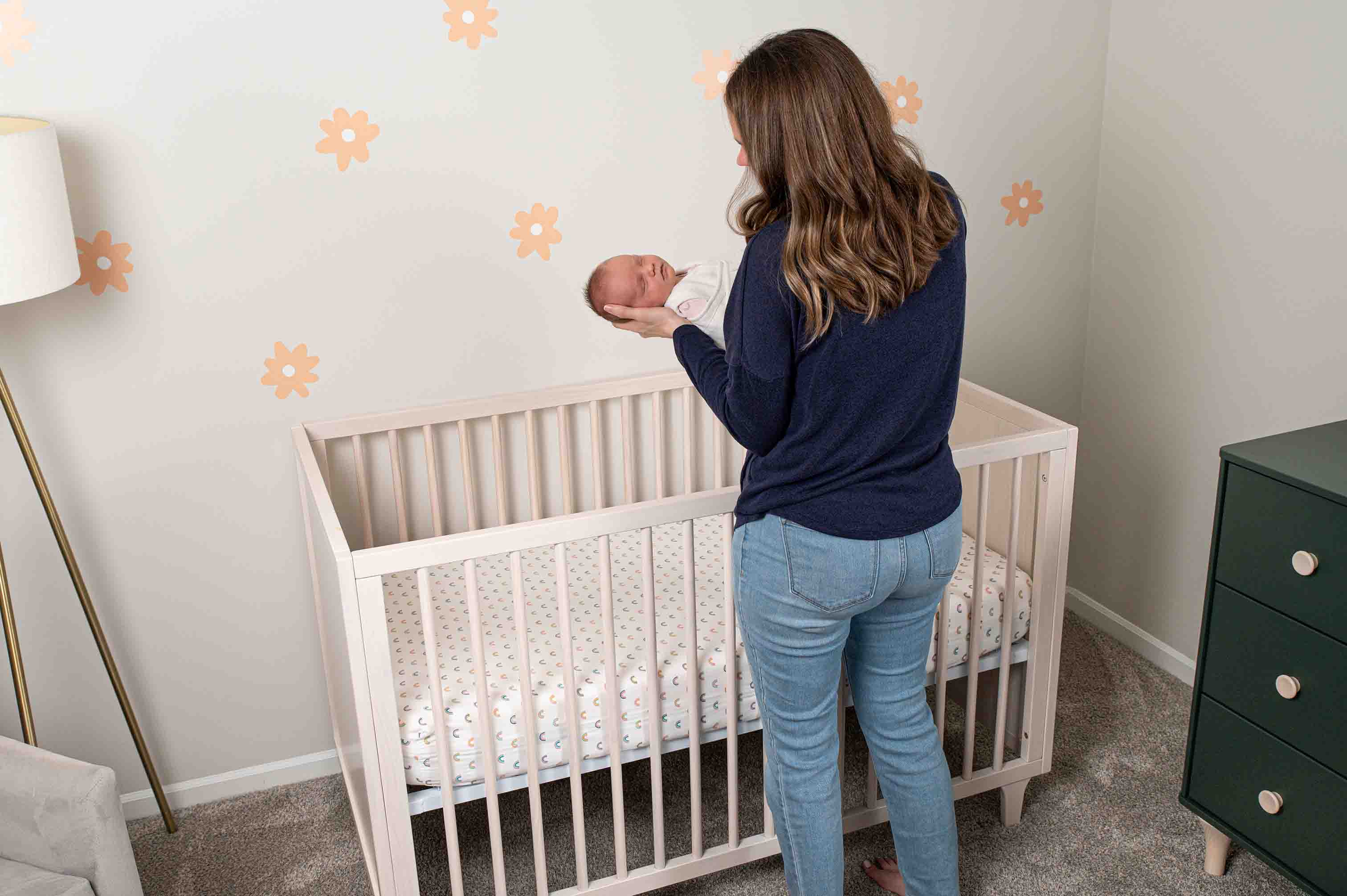A caregiver holding a baby beside a crib containing a fitted sheet and no loose bedding.