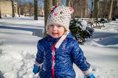 Child in blue patterned winter coat, gloves and a knitted cat hat standing against snow and trees