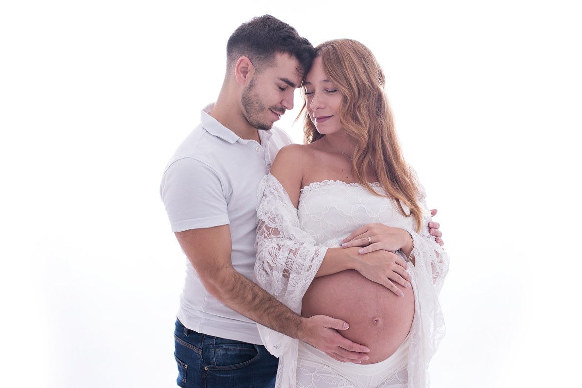 Two adults stand close with their heads touching and hands resting around a visible pregnancy bump against a bright background.