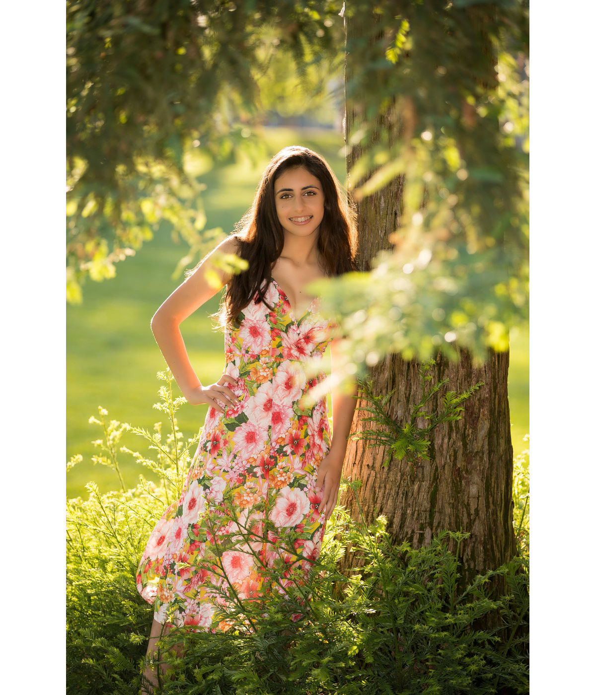 Young woman in a floral dress beside a tree, with foreground branches framing her in Jeff Cable's park portrait