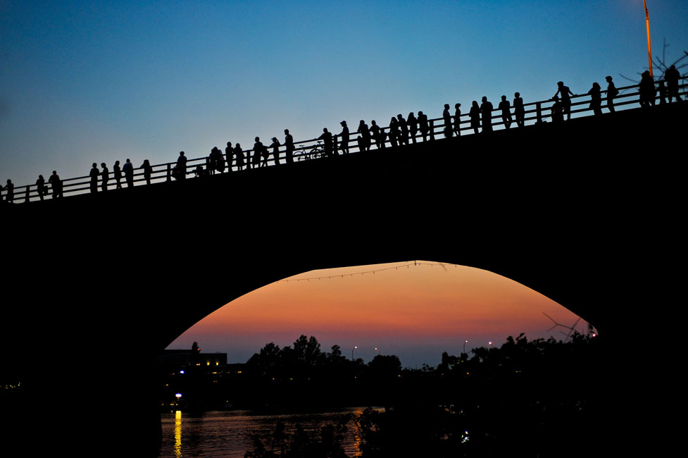 People standing along a bridge form dark outlines against a blue and orange sunset sky.