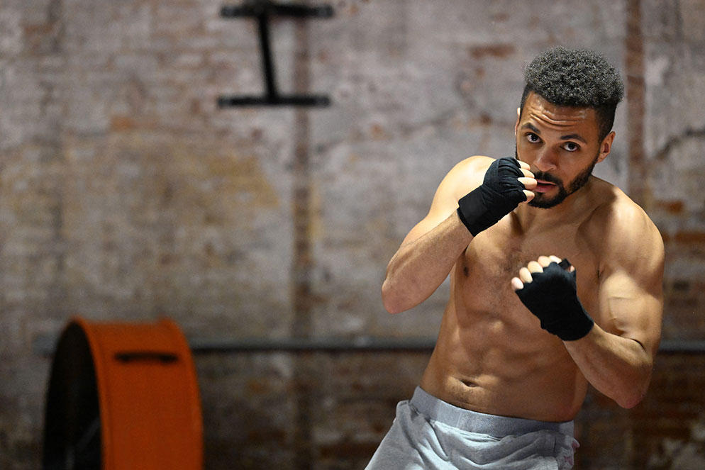 Boxer with wrapped hands raised in front of his face and chest, against a blurred indoor gym background