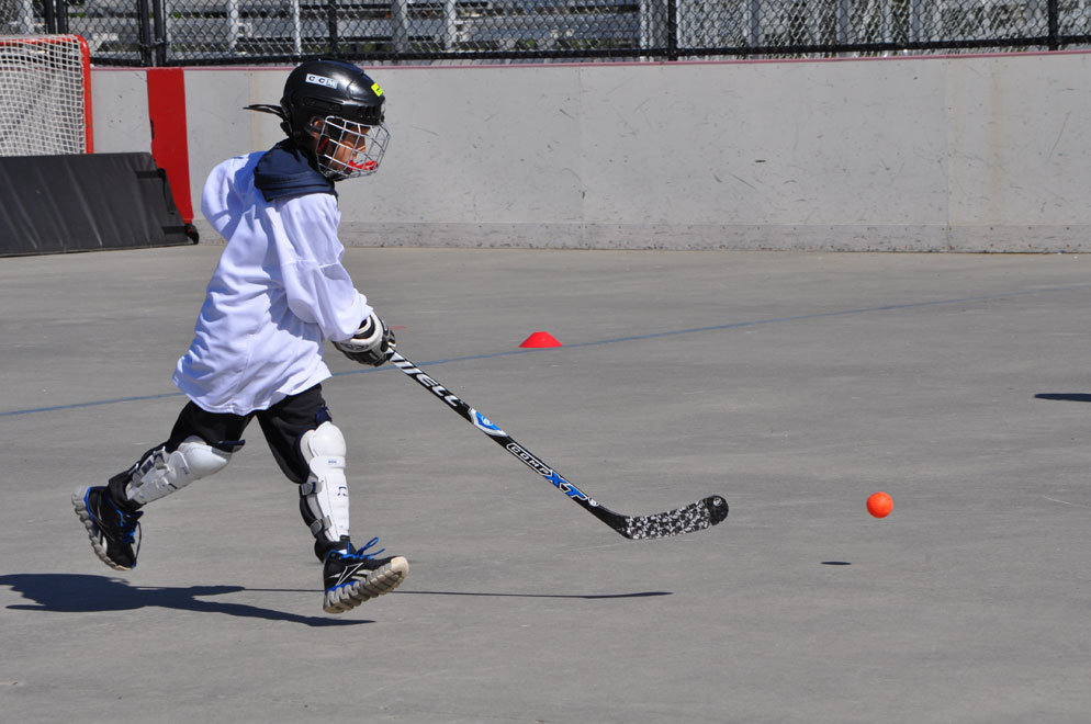 Helmeted hockey player running toward an orange ball on an outdoor paved rink, with open space ahead