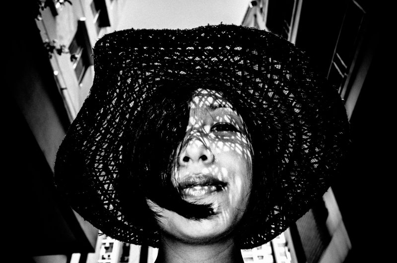 Black-and-white portrait by Eric Kim of Cindy looking upward under a woven hat, with patterned shadows across her face